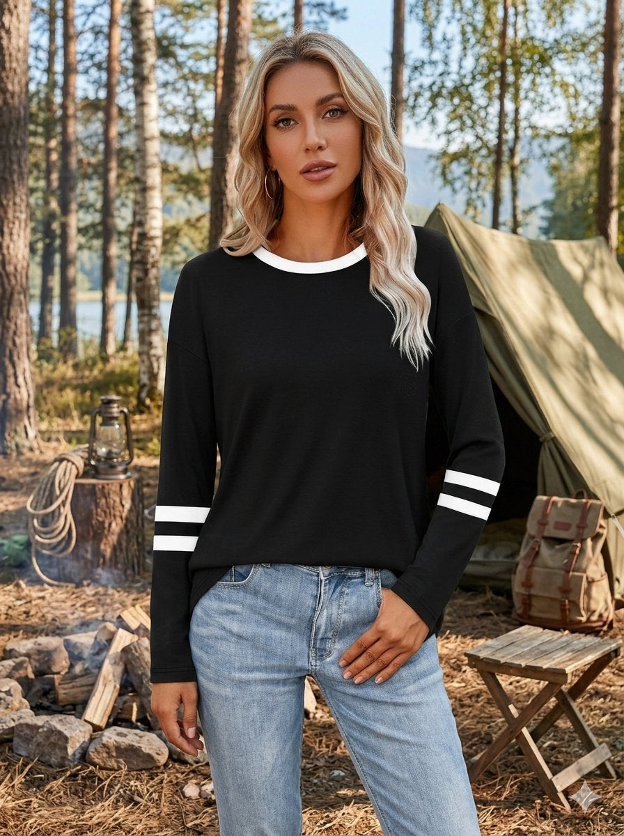A woman stands outdoors in a campsite setting wearing a black long-sleeve top with white trim at the crew neck and white stripes on the sleeves, paired with light blue jeans.