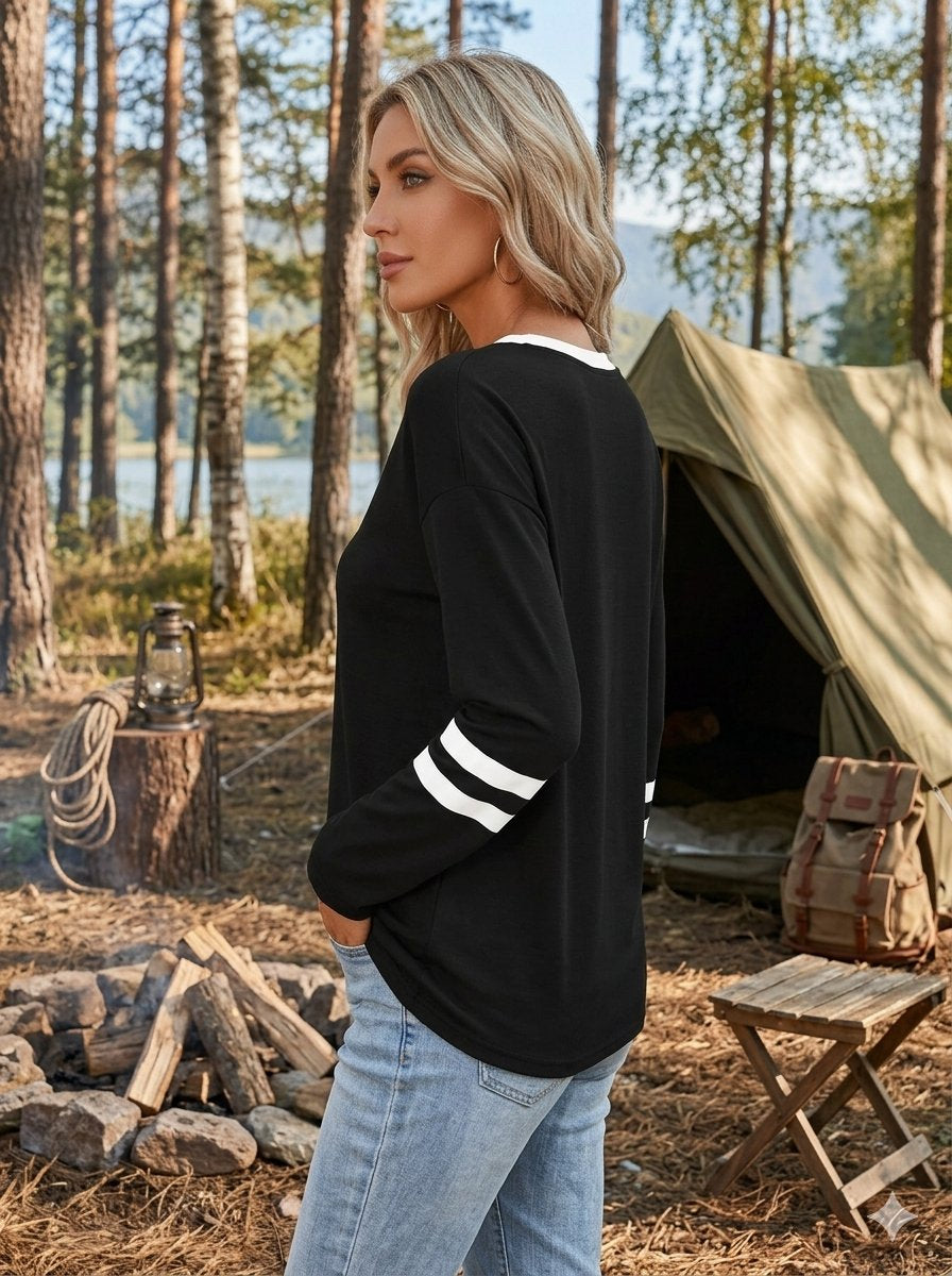 A woman stands outdoors in a campsite setting wearing a black long-sleeve top with white stripes on the sleeves and light blue jeans. Trees, a tent, logs, and camping gear are visible in the background.