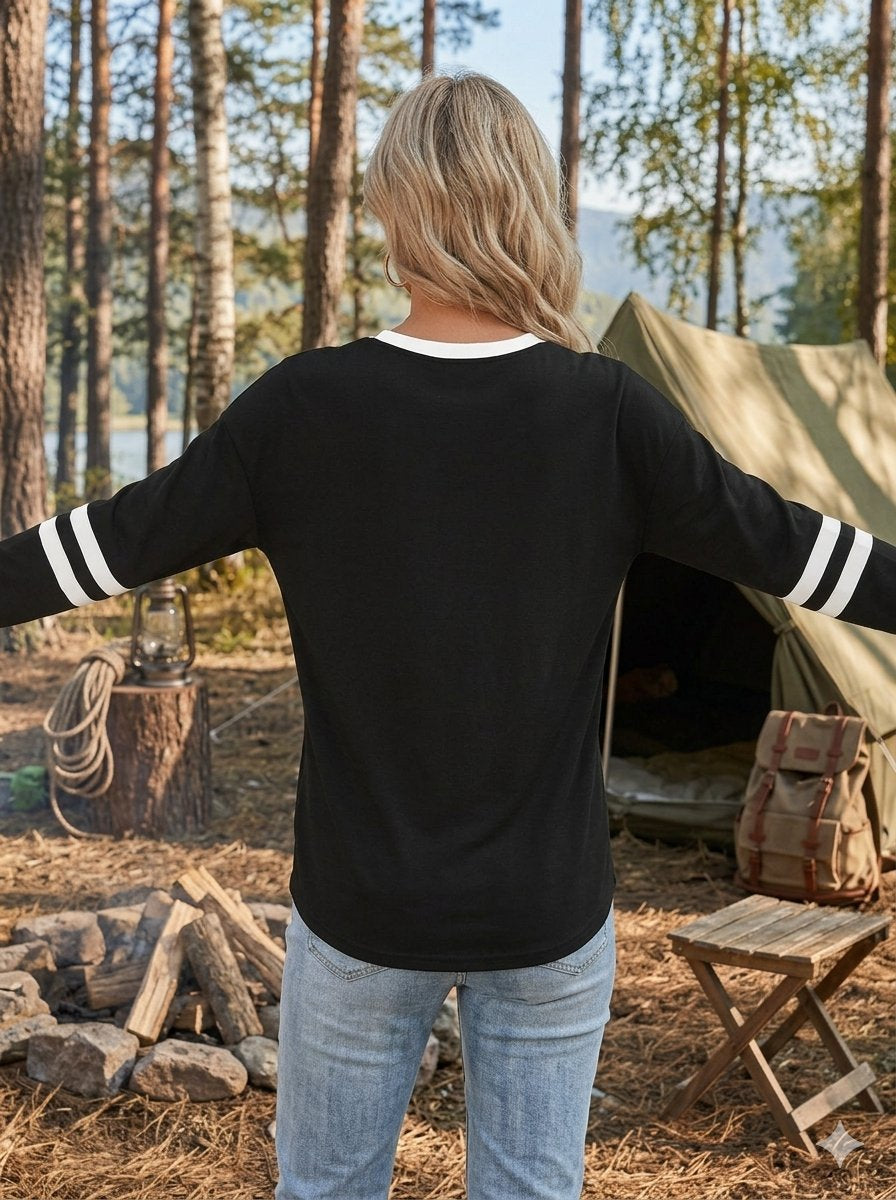 A woman stands with her back to the camera in a forest campsite setting, wearing a black long-sleeve shirt with white stripes on the sleeves and light blue jeans.