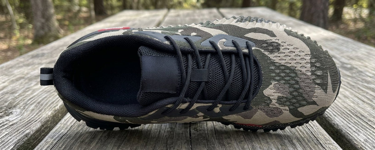A single athletic shoe with a black and camouflage pattern upper, black laces, and a black sole, resting on a weathered wooden table with a blurred forest background.