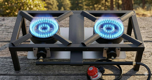 A black two-burner gas stove with blue flames lit, resting on a weathered wooden deck outdoors. A red and black gas regulator is visible in the foreground. big green bear biggreenbear.com