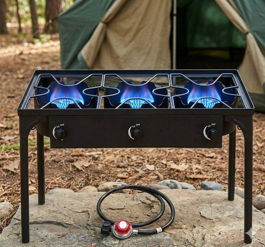 A black 3-burner camping gas stove with blue flames lit, set outdoors on a stone surface with a coiled gas hose in front and a tent blurred in the background. big green bear biggreenbear.com