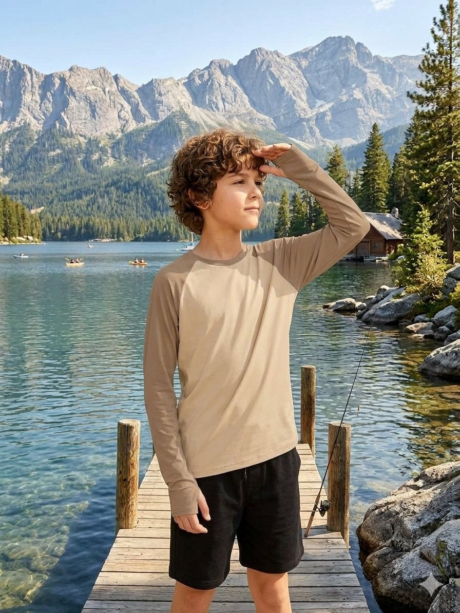 A young boy wearing a beige long-sleeve crewneck shirt and black shorts stands on a wooden dock by a scenic mountain lake, looking up toward the mountains.
