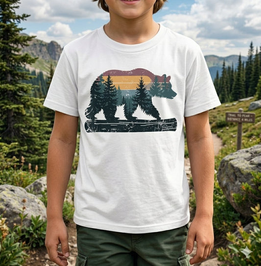 A child wearing a white graphic t-shirt with a bear and forest design, standing outdoors in a mountain landscape. big green bear biggreenbear.com