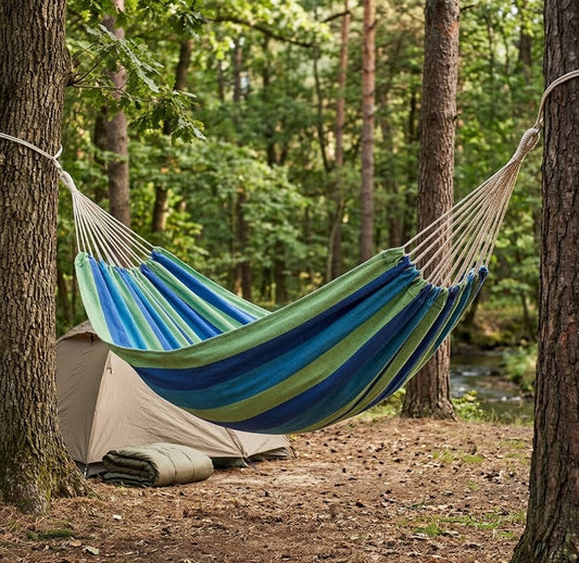 A striped camping hammock in shades of blue, green, and navy hangs between two trees in a forest setting, with a beige tent and rolled sleeping bag below. big green bear biggreenbear.com