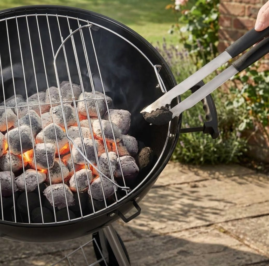 A close-up of a black round charcoal grill with glowing coals under a metal grate, with tongs holding a piece of charcoal in an outdoor backyard setting.