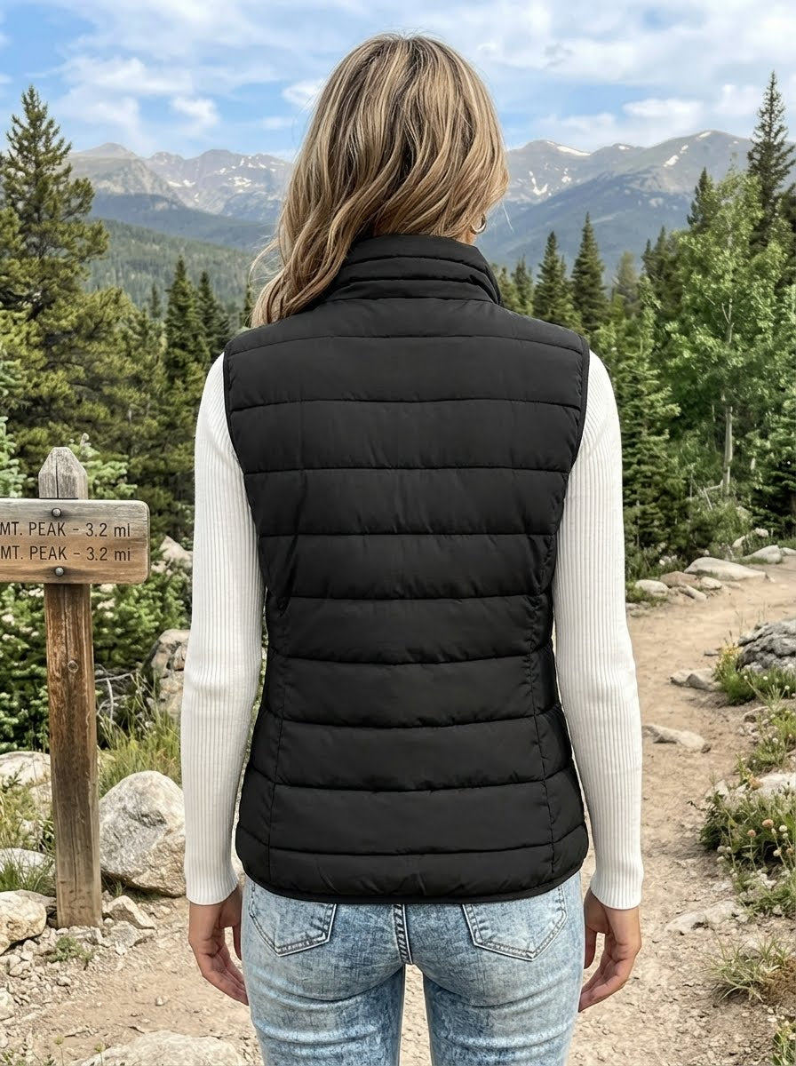 A woman viewed from behind wearing a black quilted puffer vest over a cream ribbed long-sleeve shirt and blue jeans, standing on a mountain trail with pine trees and mountains in the background.