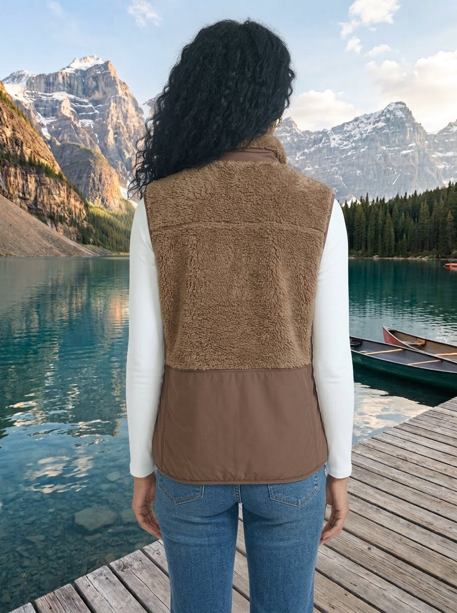 A person standing on a wooden dock by a mountain lake, viewed from the back, wearing a brown color-block fleece vest over a white long-sleeve shirt and blue jeans.