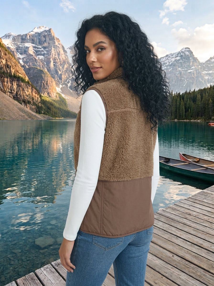 A woman with long curly black hair stands on a wooden dock by a blue lake with mountains in the background, wearing a brown color-block fleece vest over a white long-sleeve top and blue jeans.