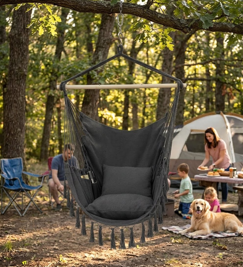 A dark gray fabric sling hammock chair with a wooden spreader bar and tassel fringe, hanging from a tree in a wooded outdoor campsite setting with people, a tent, and a dog in the background.