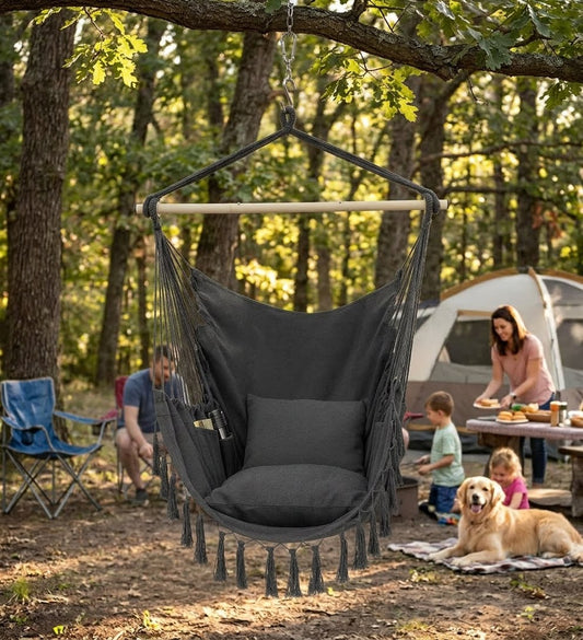 A dark gray fabric sling hammock chair with a wooden spreader bar and tassel fringe, hanging from a tree in a wooded outdoor campsite setting with people, a tent, and a dog in the background.