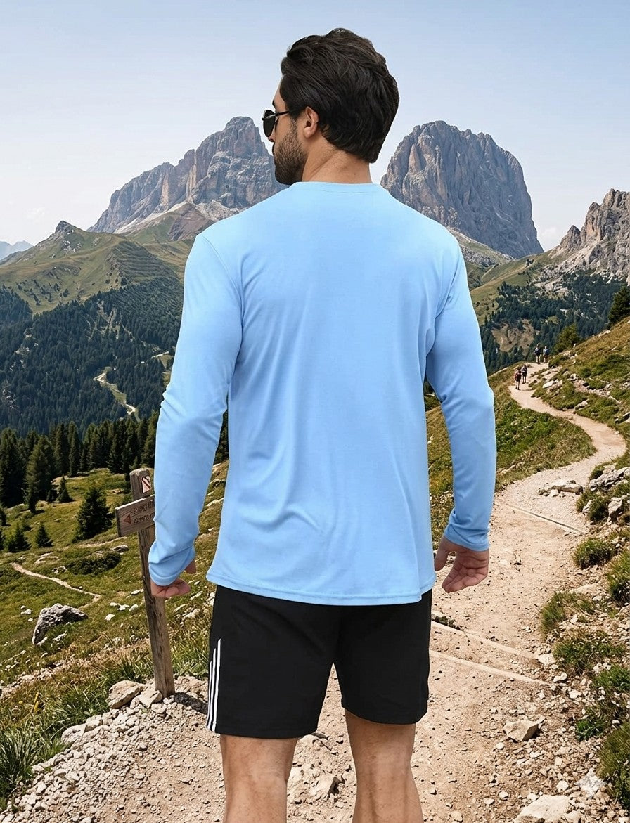 Man in a blue long-sleeve shirt and black shorts walking on a mountain trail with mountains in the background.